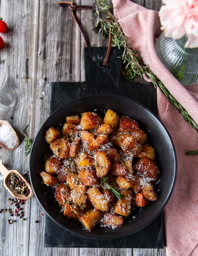 A plate of crispy potatoes on a. black marble surrounded by whole herbs and peppercorns