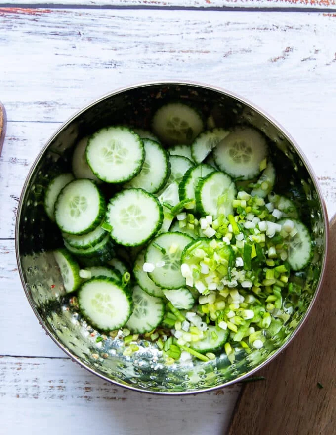 The sliced cucumbers and scallions added into the salad bowl of the dressing
