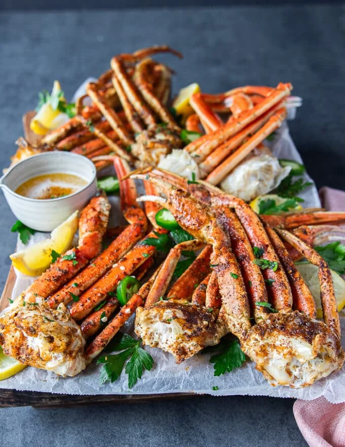 side view of a plate of cooked crab legs 4 different ways on a board surrounde dby melted butter in a bowl and lemon wedges