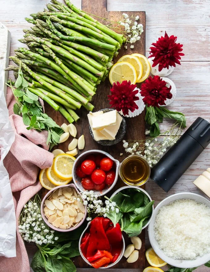 remaining ingredients for the lamb chops including the asparagus to air fry, some roasted red peppers in a jar, some garlic cloves, some whole basil leaves, grated parmesan cheese in a bowl, some olive oil and butter in a bowl, some almonds, some lemon juice in bowls , seasoning