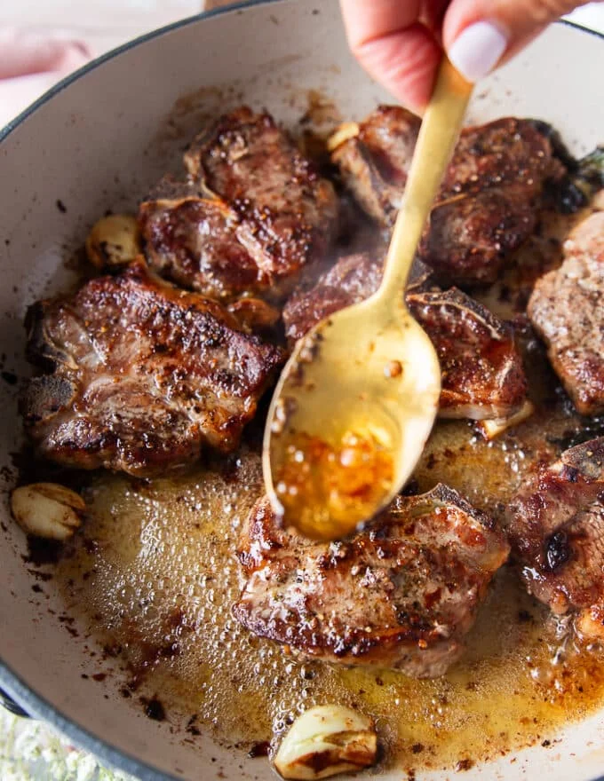 A spoon basting some butter from the skillet over the lamb chops to infuse them and cook them