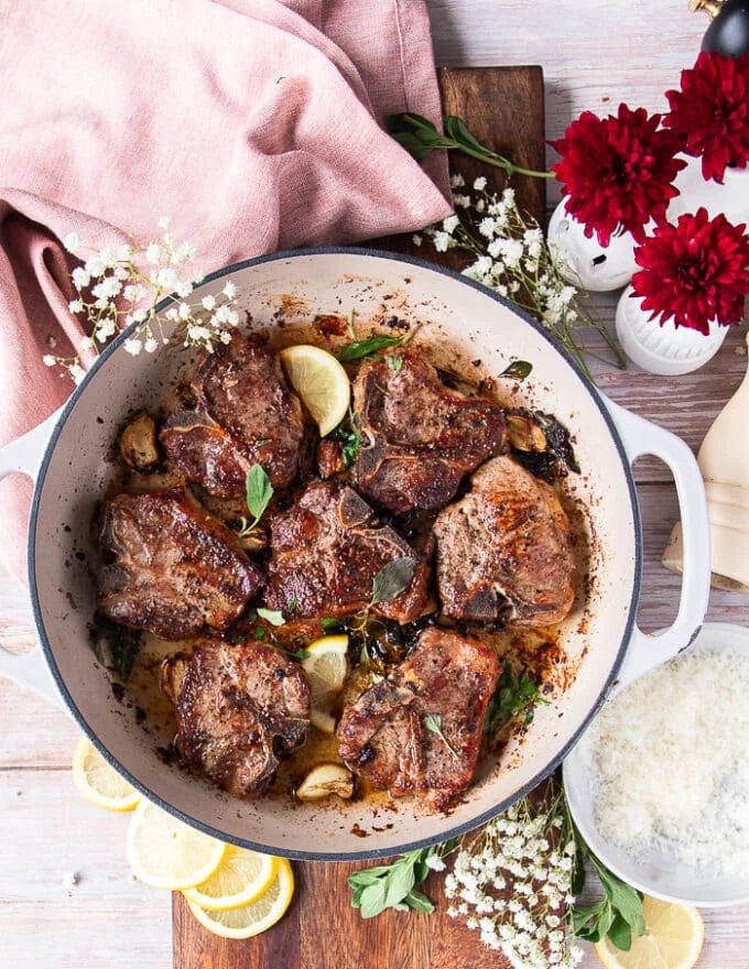 A skillet with perfectly cooked lamb chops with fresh herbs, whole garlic cloves and lemon slices
