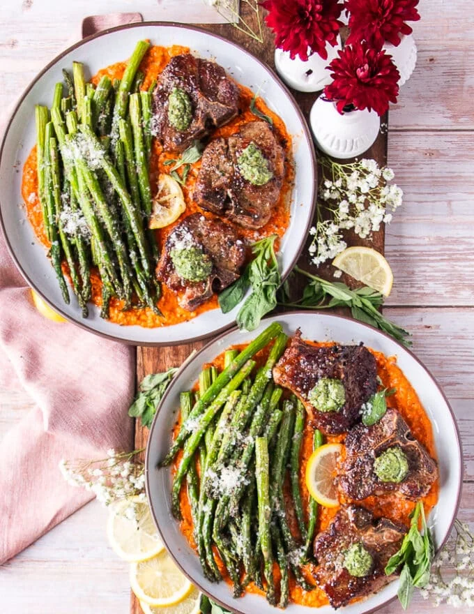 two plates of cooked lamb chops, asparagus, romesco sauce, and basil pesto
