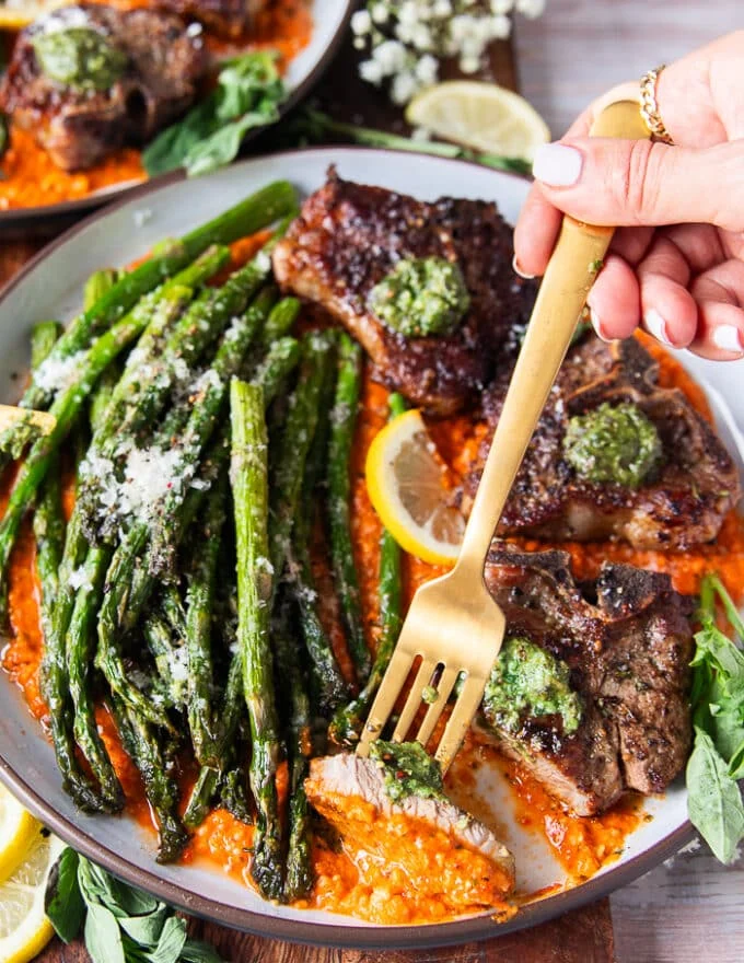 A hand holding a fork with a cut up piece of lamb chops, some pesto and romesco sauce on top
