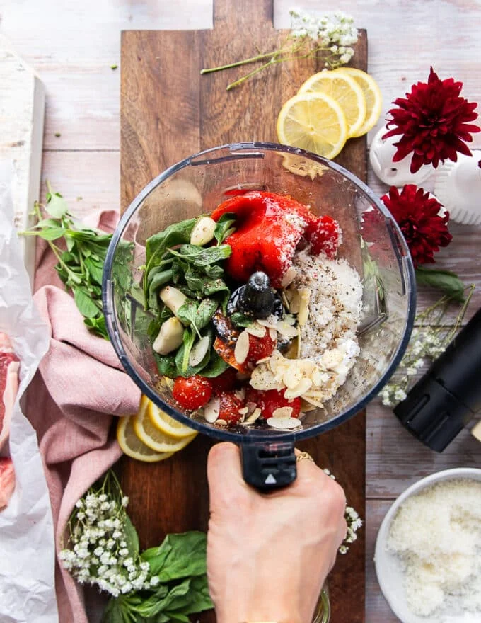 A food processor with all the romesco sauce ingredients added in one go including roasted red peppers, cherry tomatoes, almonds, lemon juice, fresh basil, olive oil, garlic, parmesan cheese and lemon juice