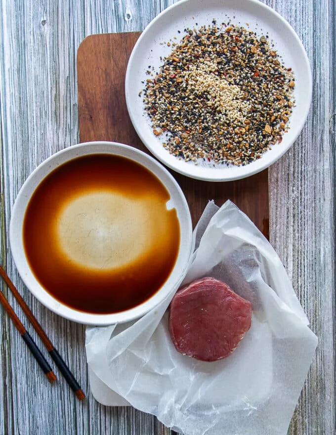 ingredients for seared ahi tuna including a piece of ahi tuna, a bowl of soy sauce, and a plate of sesame seeds, dried onion flakes, oregano , chilli powder and coarse pepper for the crust