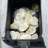 The thinly sliced and dried potatoes in the basket of the air fryer sprayed with some oil spray