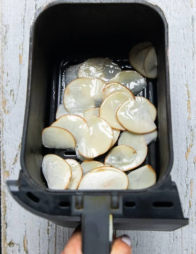 The thinly sliced and dried potatoes in the basket of the air fryer sprayed with some oil spray