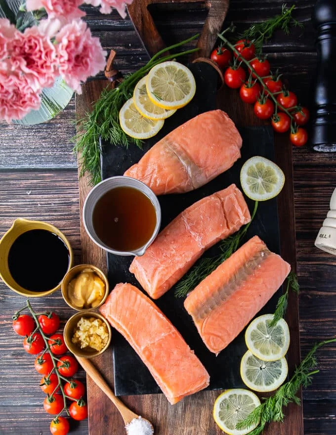 ingredients for maple glazed salmon on a board such a as salmon fillets, a bowl of maple syrup, a bowl of soy sauce, a bowl of mustard, a bowl of minced garlic, salt and pepper mills and olive oil