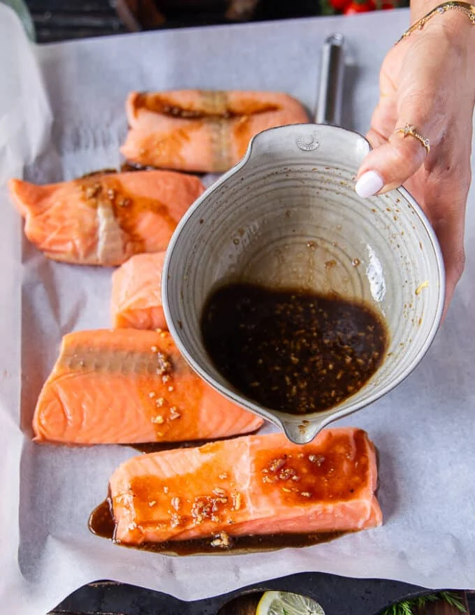 A hand pouring half of the maple glaze form the bowl on to the salmon fillets on a baking sheet