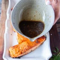 A hand pouring the remaining maple glaze over the baked salmon to finish the dish
