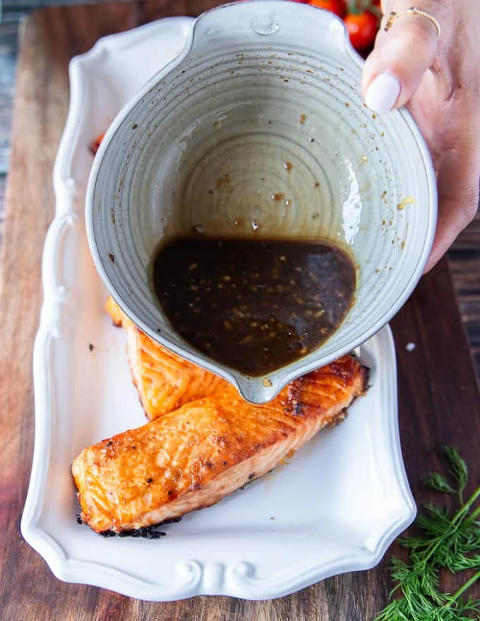 A hand pouring the remaining maple glaze over the baked salmon to finish the dish