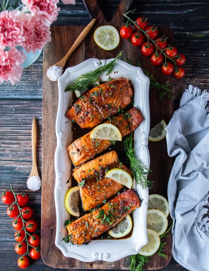 A full plate of 4 pieces of maple glazed salmopn fillets surrounded by lemon slices, some fresh herbs and drenched in maple glaze