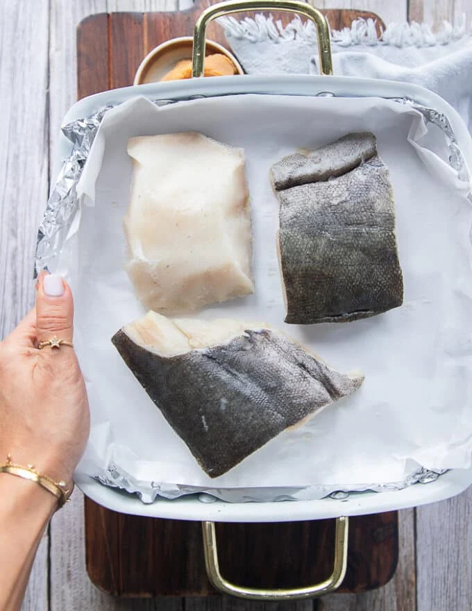 A hand holding a tray of black cod showing what the fish is and how black the skin is and how thick the fillet is.