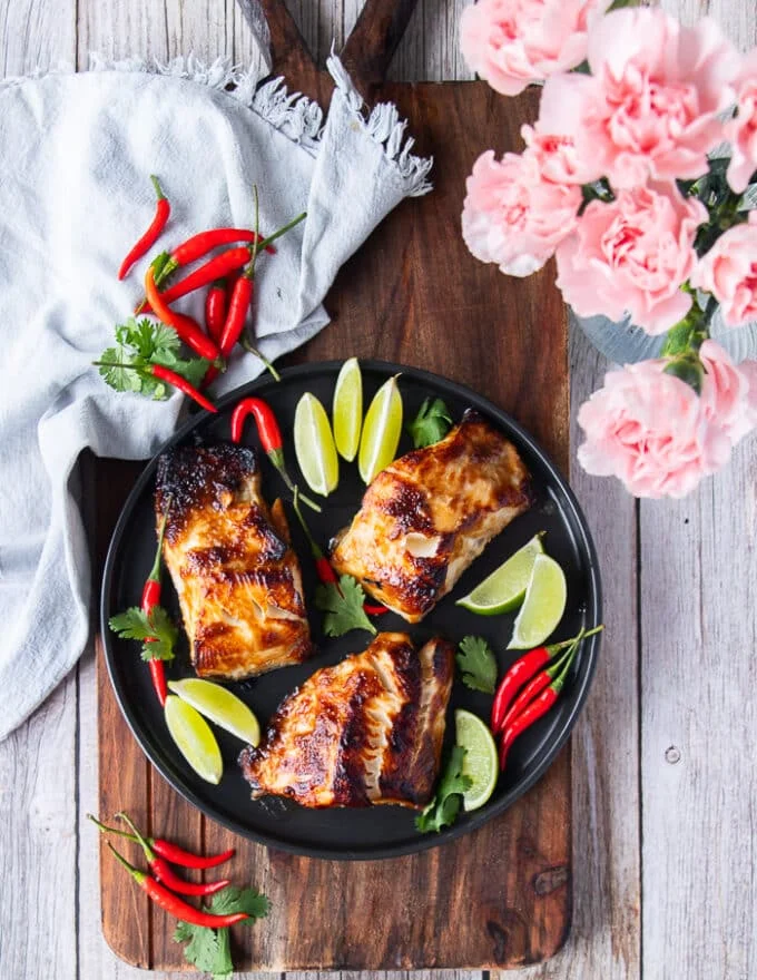 A full plate of miso black cod with lime wedges and a tea towel on a kitchen table and some flowers on a vase