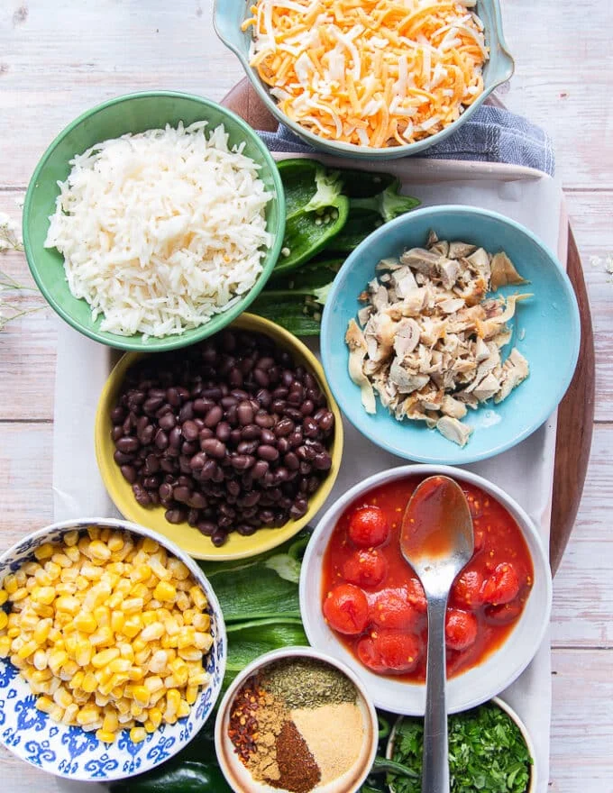 stuffed poblano peppers ingredients including a bowl of cooked chicken , a bowl of cooked rice, a bowl of beans, a bowl of corn, a bowl of tomatoes, a bowl of cheese, some cilantro, spice in a bowl, the poblano peppers
