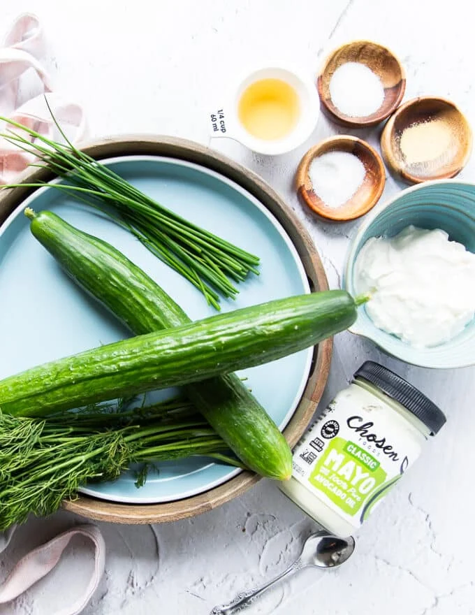 Ingredients for creamy cucumber salad including two large cucumebrs, a bowl of sour cream a few tablespoons mayo, some garlic powder and seasoning, lemon juice, a bunch of fresh dill
