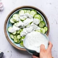 A hand pouring the creamy dressing over the cucumber, dill and onions