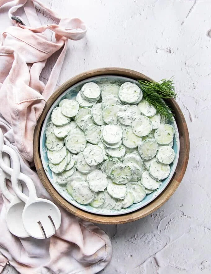 Top view of final creamy cucumber salad surrounded by salad spoons and a kitchen towel garnished with lots of fresh dill