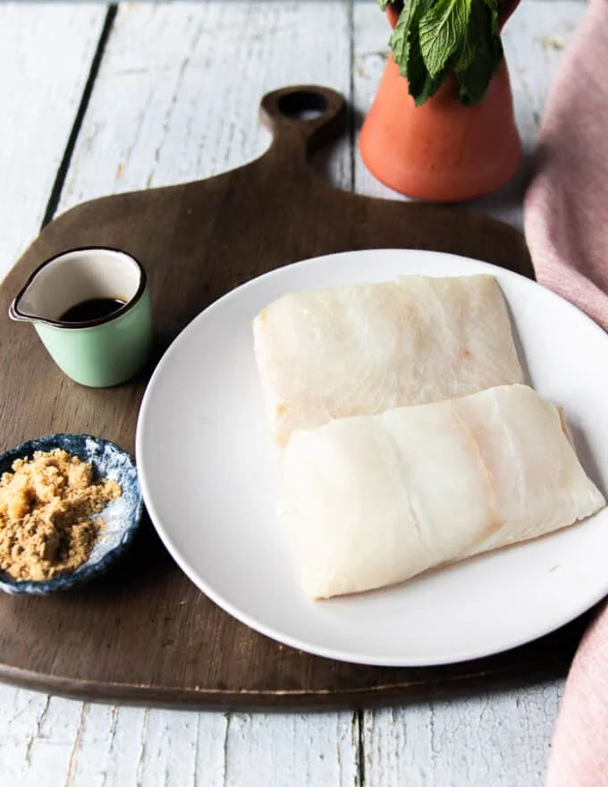 Ingredients to make grilled halibut on a board including two halibut fillets, some soy sauce in a bowl, dried ginger in a bowl and light brown sugar in another bowl