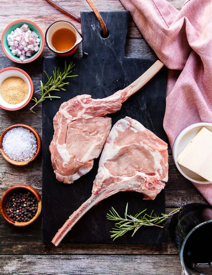 Ingredients for grilled veal chops on a board including seasoning in small bowls, some balsamic vinegar, some shallots minced, butter in a bowl, fresh rosemary sprigs, honey in a small bowl and two veal chops