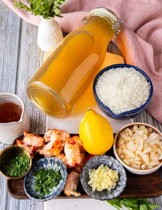 ingredients needed to make lobster risotto in a board including a jar of lobster stock, a cup of arborio rice, some butter poached lobster in a bowl, some lemon zest, some minced garlic in a bowl, some minced shallots in a bowl some butter from the lobster poaching