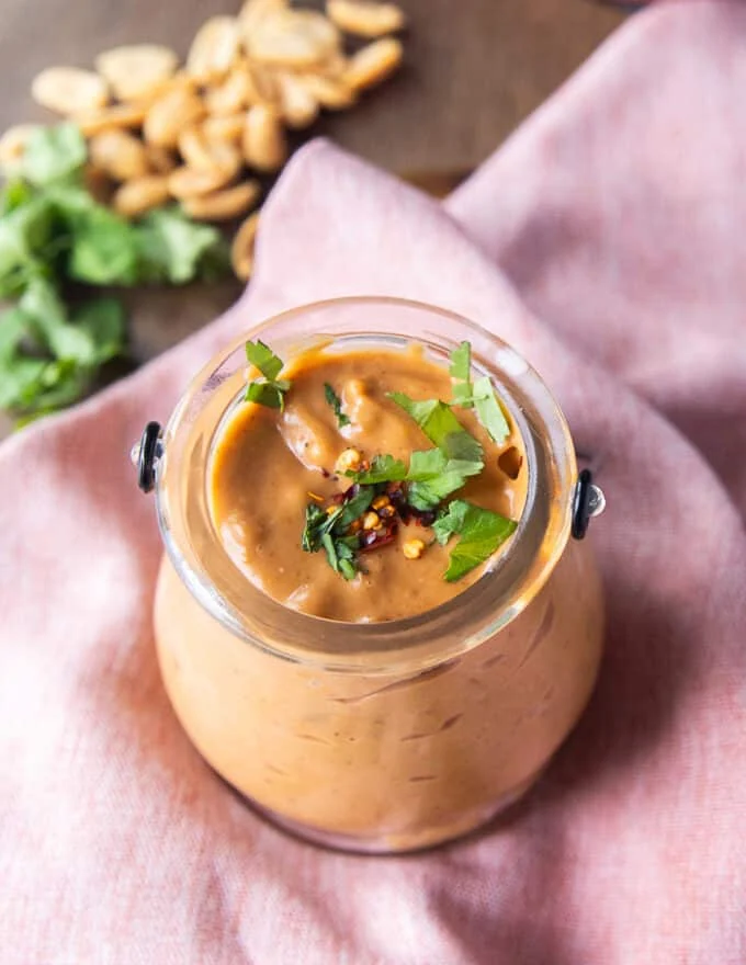 A jar of satay sauce garnished with crushed peanuts, cilantro and chilli flakes