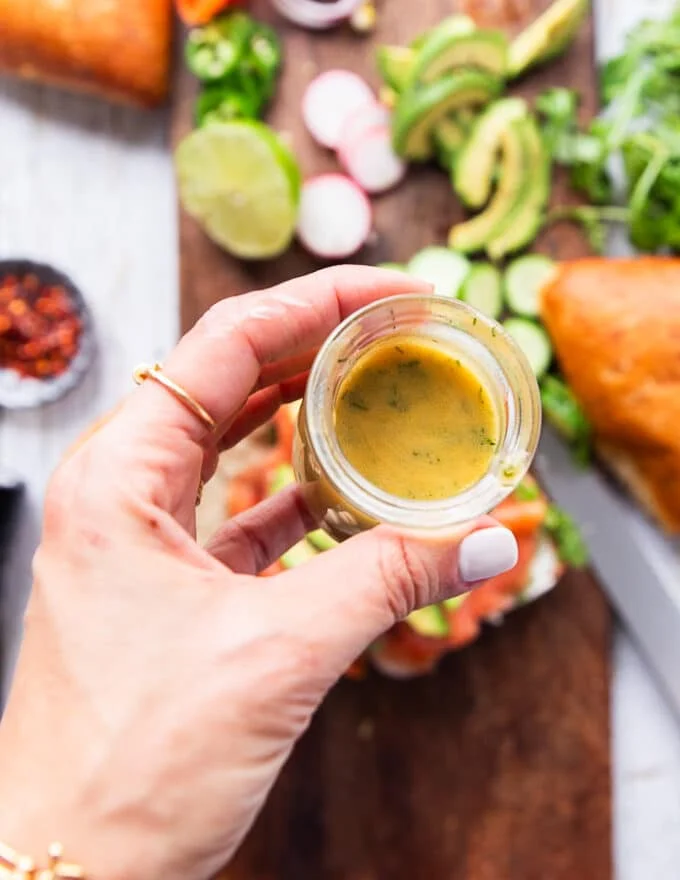 A hand holding the dill mustard sauce and ready to drizzle it over the smoked salmon to finish the sandwich