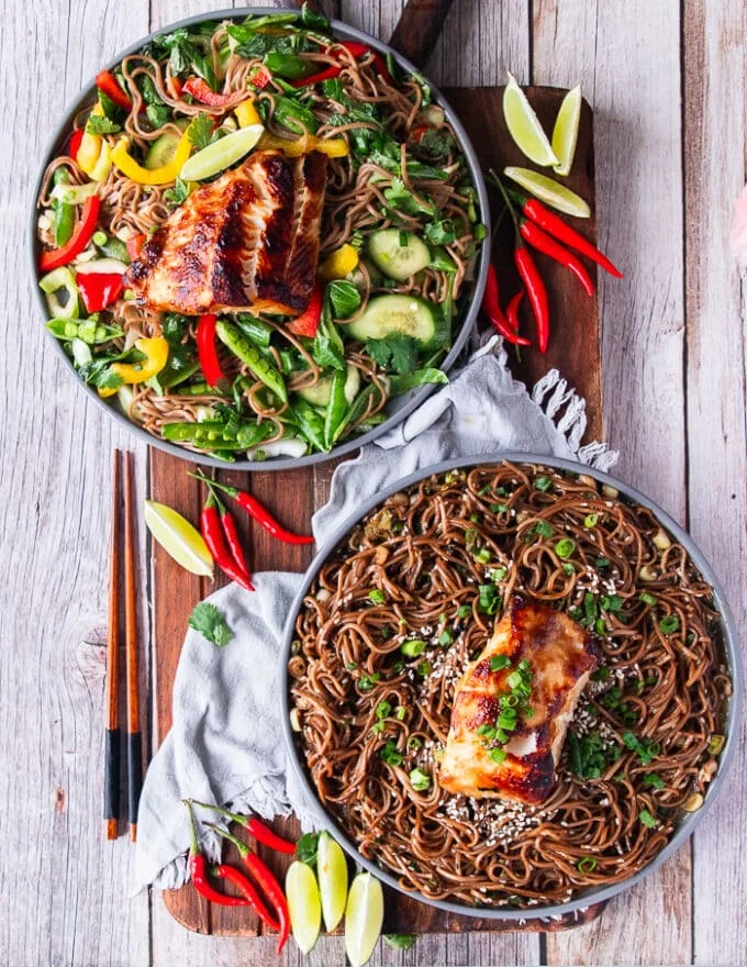 two plates of soba noodles, one served warm and the other cold with veggies