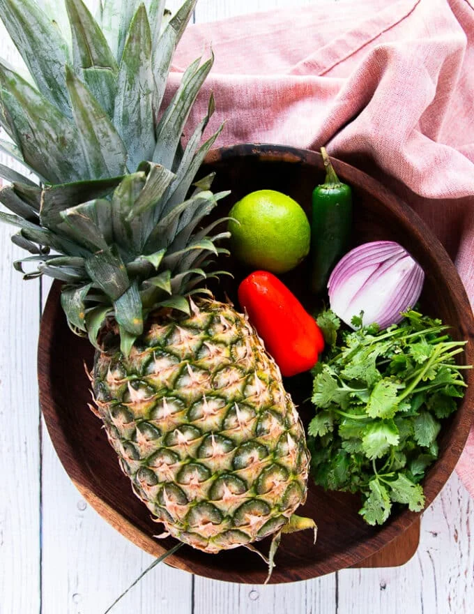 A large wooden bowl with pineapple salsa ingredients including a large fresh pineapple, a bunch of cilantro, a lime, a red onion, a bell pepper, and salt