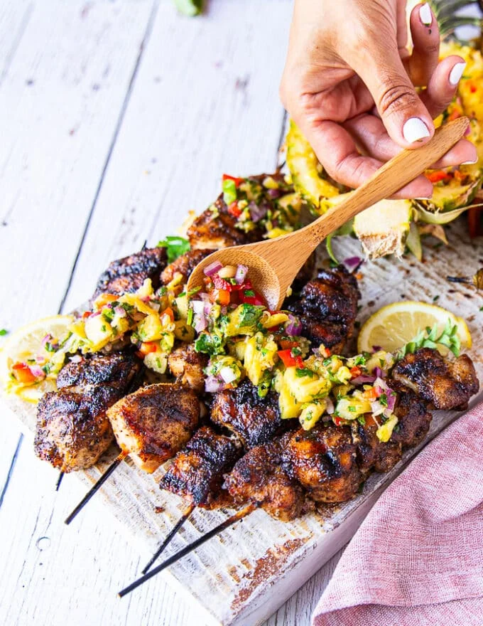 A hand scooping up some pineapple salsa over grilled chicken skewers showing one of the many ways to use it other than dipping chips