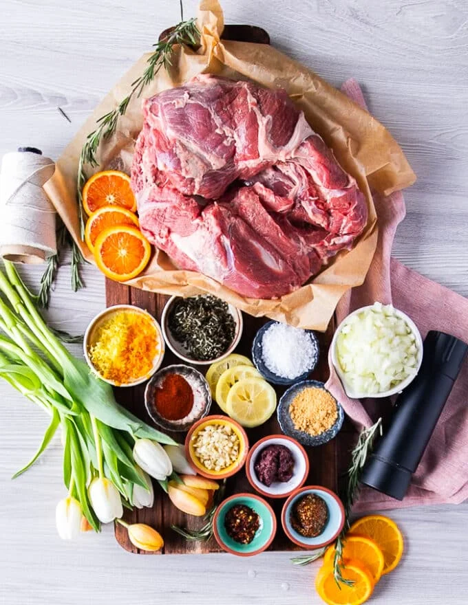 All ingredients needed for roast boneless leg of lamb including boneless leg of lamb, some citrus zests, some onions, mustard, spices in separate bowls, some fresh rosemary, some garlic, lots of olive oil.