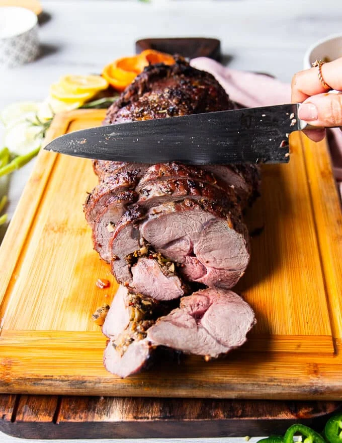 A hand holding a knife and slowly slicing the perfect boneless leg of lamb showing the first slice how perfectly cooked and flavored it looks