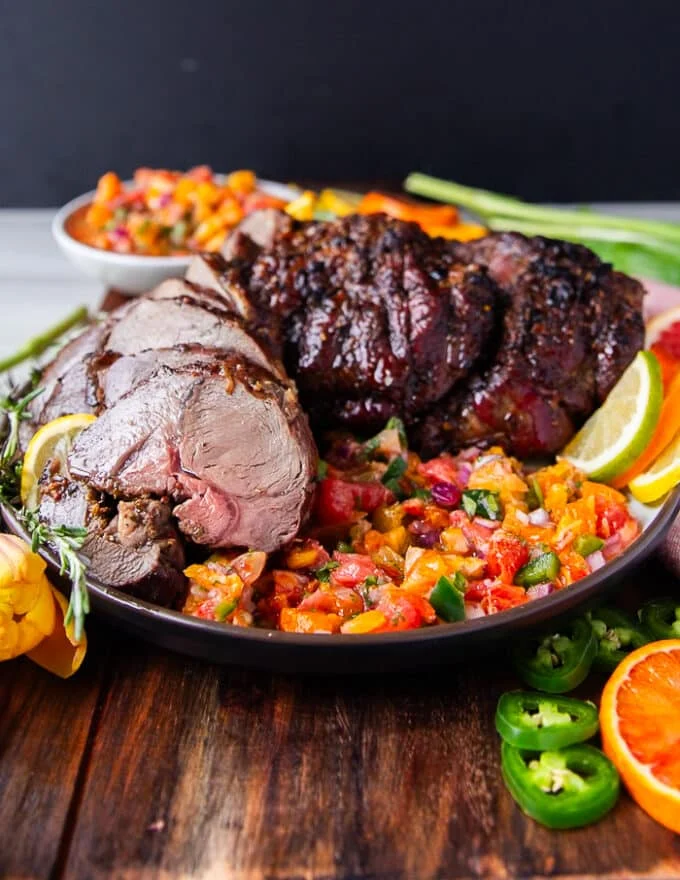 A side view of a roasted leg of lamb boneless and served on a platter with fresh herbs and citrus salsa