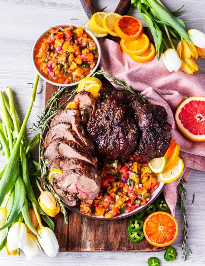 A whole roasted leg of boneless lamb sliced on a platter drizzled with come citrus salsa and garnisheed with lots of fresh rosemary and citrus slices