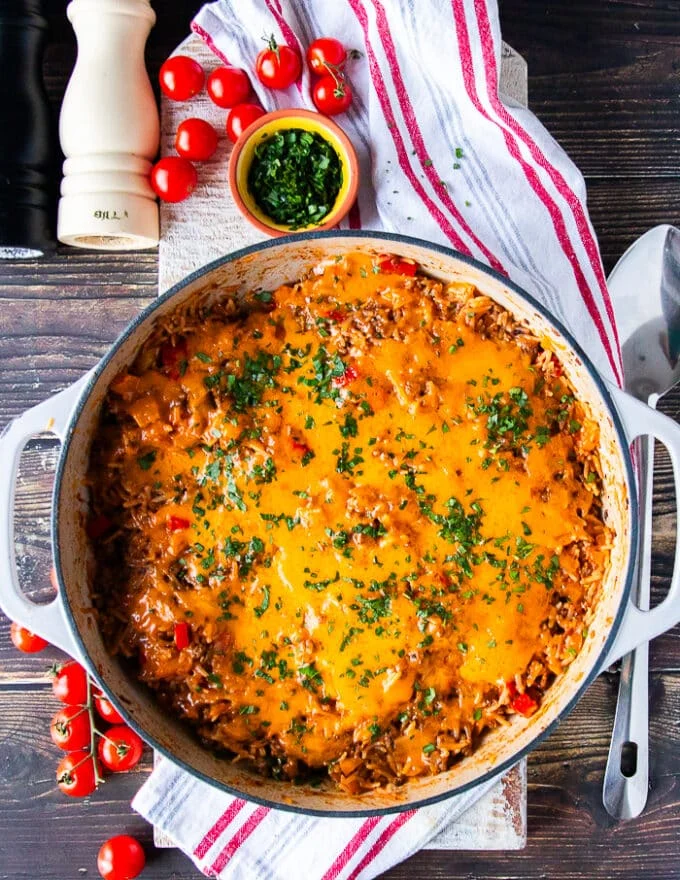 Top view of stuffed pepper casserole surrounded by a kitchen towel some baby tomatoes on the vine. fresh herbs and a bowl of chilli flakes