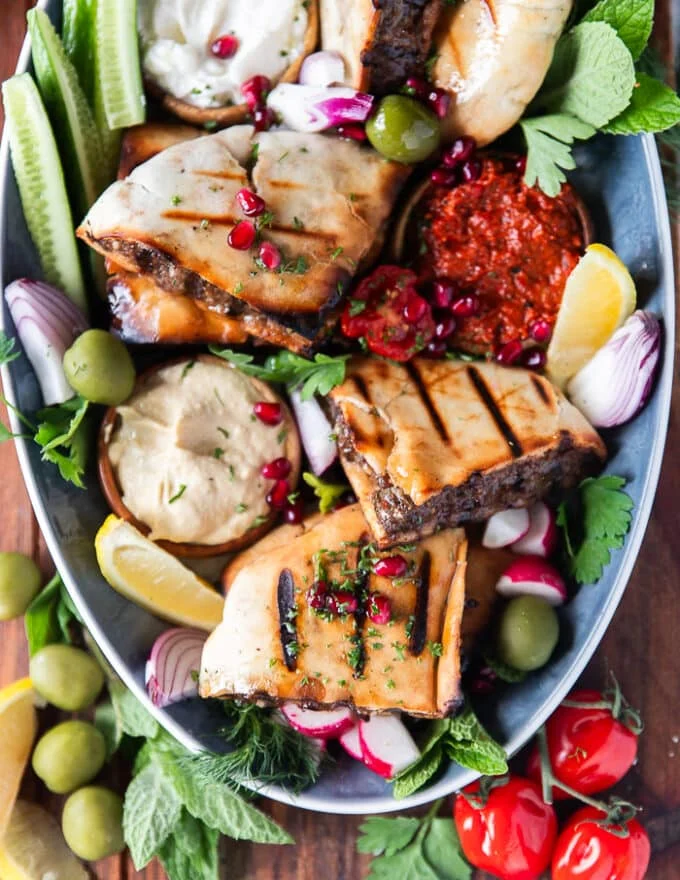 A plate of Arayes grilled and arranged surrounded by hummus dips, ypgrut dips, and muhammara, some lemon wedges, some chilli flakes