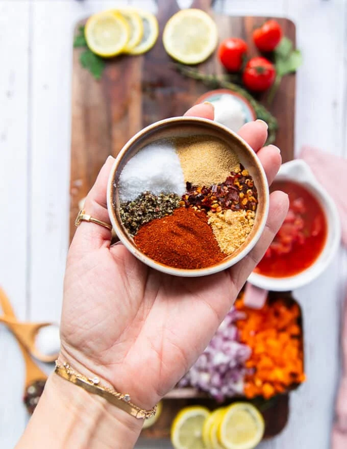 A hand holding a plate of homemade cajun seasoning ingredients including some smoked paprika, some salt, pepper , oregano, some garlic and onion powders