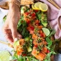 A hand holding a piece of salmon carpaccio with pita chips showing closeup of the salmon, the dressing and the capers
