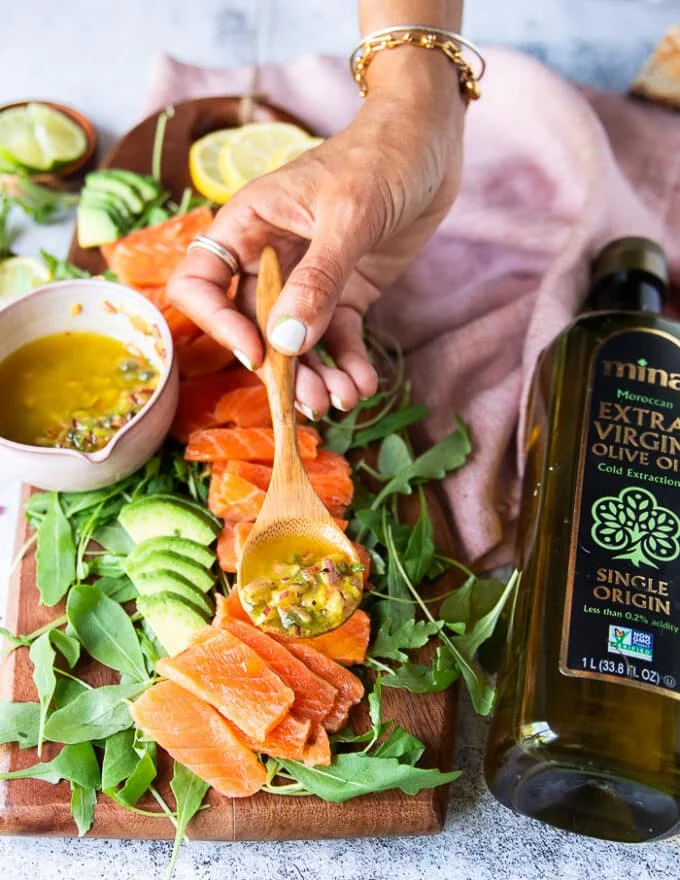 A hand holding a spoonful of the vinaigrette and dressing the salmon slices with it to marinate and season the salmon carpaccio