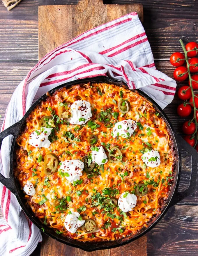 A finished skillet of taco spaghetti on a wooden board with baby tomatoes around it, some chilli flakes in a spoon and red and white tea towel, some herbs