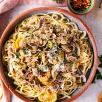 A bowl of clam pasta in a clay bowl surrounded by small dishes of herbs, lemon slices and a bowl of chilli flakes.