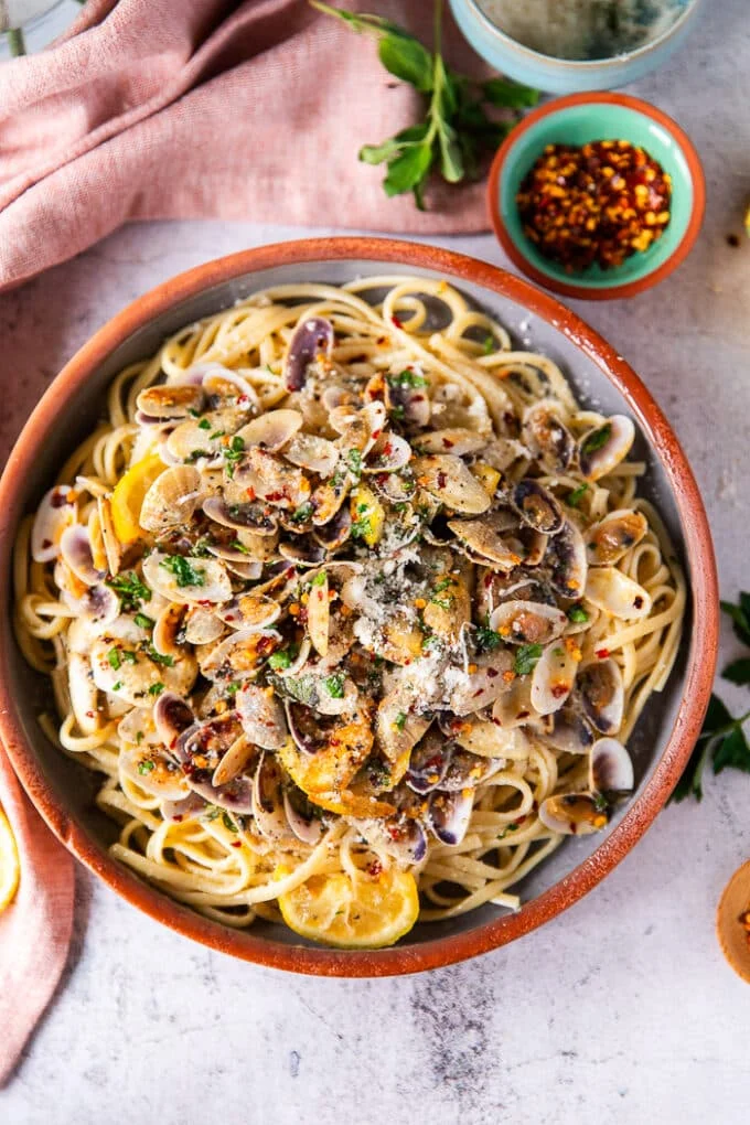 A bowl of clam pasta in a clay bowl surrounded by small dishes of herbs, lemon slices and a bowl of chilli flakes.