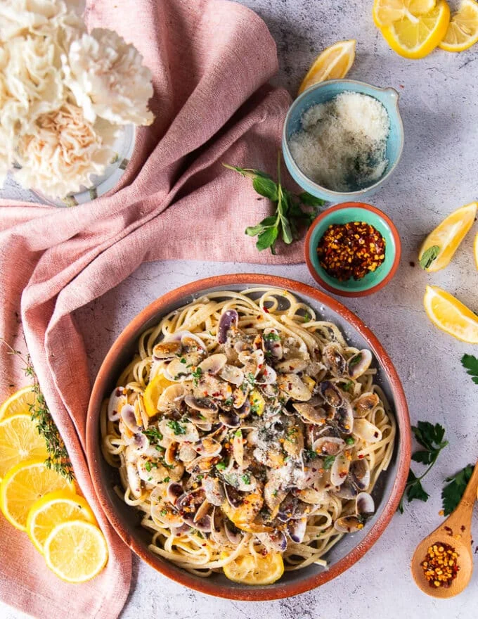 fully set up clam pasta dish surrounded by flowers and some lemon slices, a tea towel, some spices and a bowl of fresh Parmesan cheese