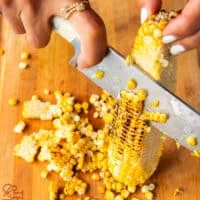 a hand holding a corn on the cob vertically on a wooden board and then slicing the corn kernels using a sharp knife