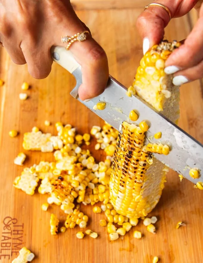 a hand holding a corn on the cob vertically on a wooden board and then slicing the corn kernels using a sharp knife
