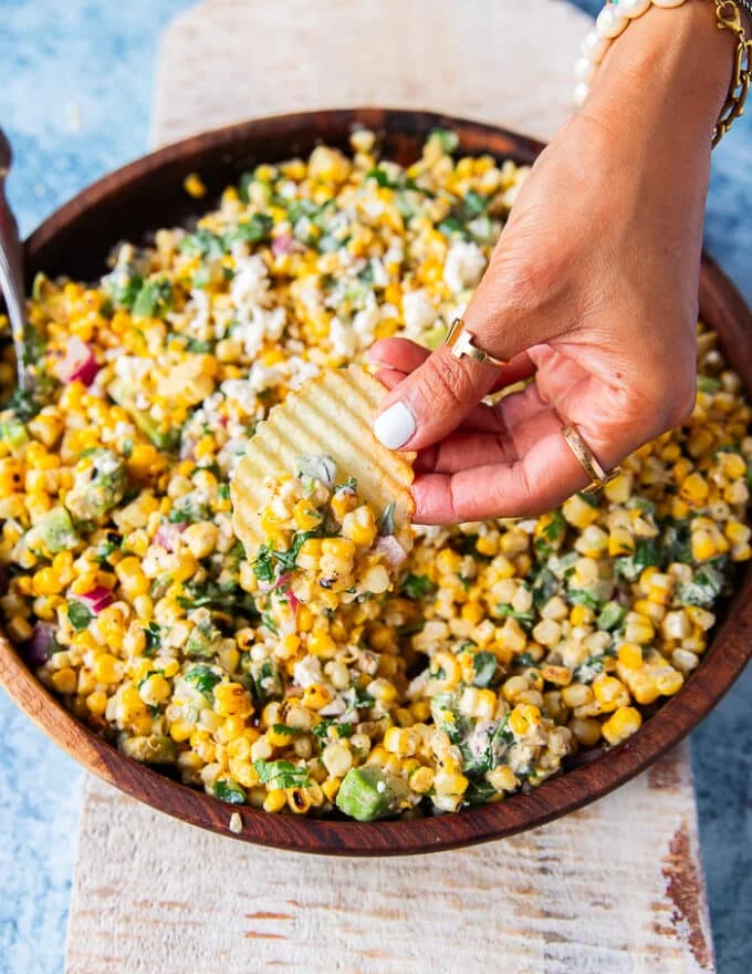A hand holding a chips and scooping some mexican corn salad with creamy cilantro lime dressing, jalapenos and avocados