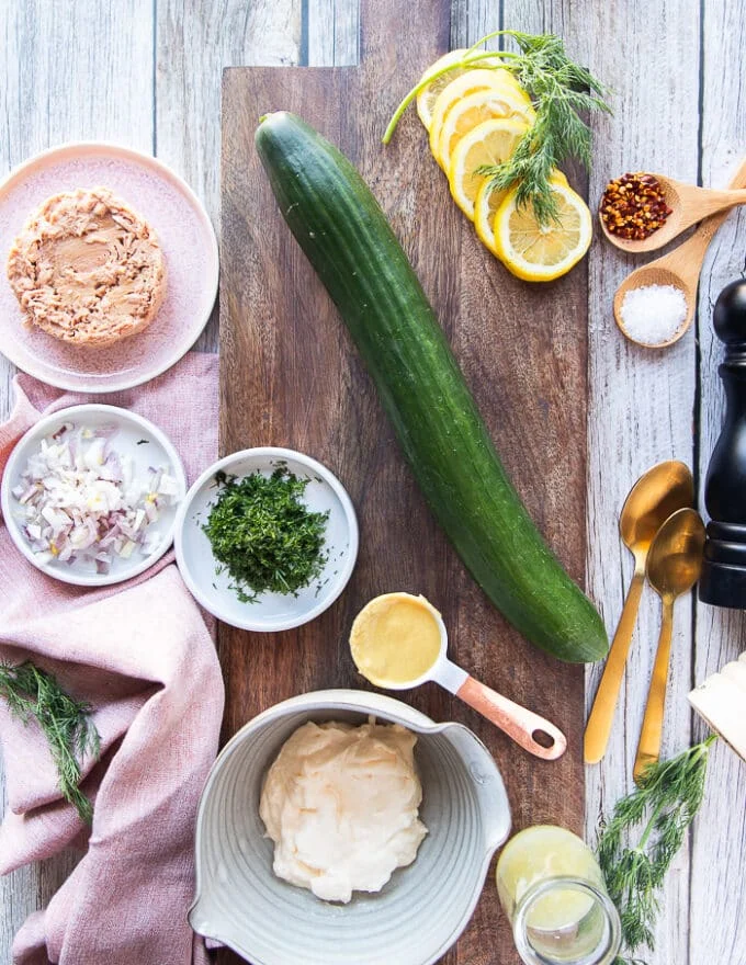 Ingredients for tuna cucumber salad including a whole cucumber, canned tuna in a bowl, fresh dill chopped, some mayo in a bowl, some spices, some onions chopped, lemon juice