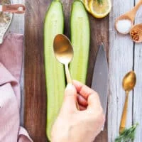 a hand holding a spoon and scooping the flesh from the sliced cucumbers