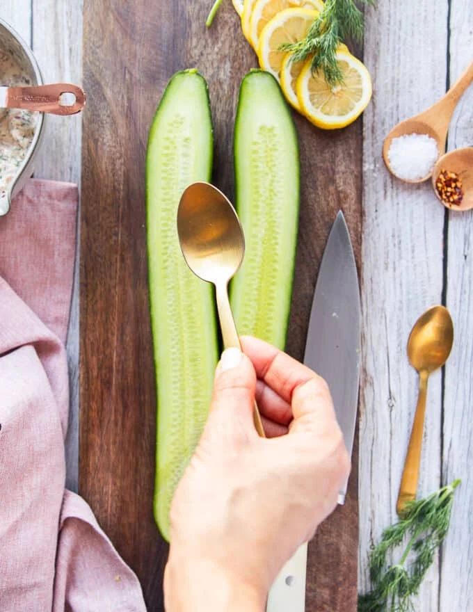 a hand holding a spoon and scooping the flesh from the sliced cucumbers